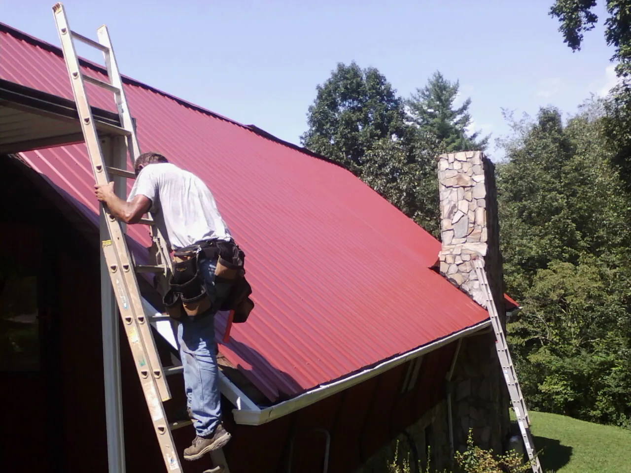 Roofer on a ladder beside a red metal roof and stone chimney — Soffit & Fascia Repair by Roofing Brothers in Klamath River